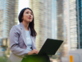 Young Asian woman looking up while using laptop, sitting at park against office buildings in the city.