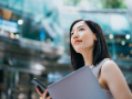 Successful and modern young Asian businesswoman carrying smartphone and laptop, commuting to work in central business district against contemporary corporate buildings in the city.