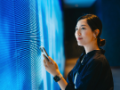 Confident young Asian woman using smartphone against blue-colored illuminated LED digital display screen, connecting to the future.