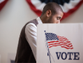 Man in white shirt and black vest standing at a voting booth.