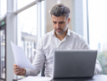 Man in white shirt with gray beard working at desk with laptop and papers in office. 