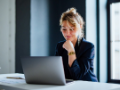 Smiling businesswoman standing at desk with cup of coffee and typing business report on a laptop computer keyboard.