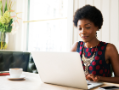 Woman with gold necklace, laptop, and coffee cup at table.