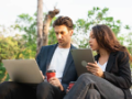 Young businessman showing content on tablet/laptop to female colleague at bench in park.