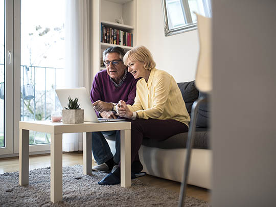 senior couple sitting on edge of couch smiling at laptop