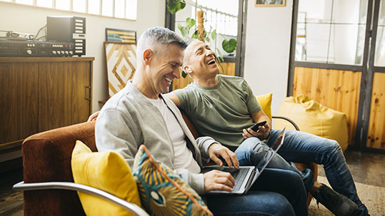 casually-dressed middle aged men sitting on couch using a laptop laughing