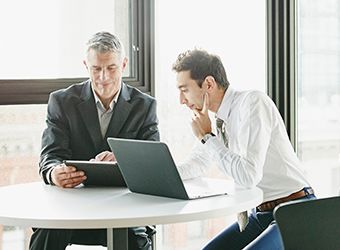 two Caucasian business men with laptop seated at table looking at tablet image